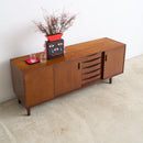 A mid-century modern wooden sideboard cabinet with a warm brown finish, featuring a vase with dried branches on top, set against a plain white wall and light concrete floor.