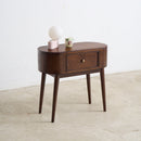 A mid-century modern dark brown wooden side table with a single drawer and tapered legs, styled with a pink lamp and a small floral mug on top, set against a plain white wall and concrete floor.