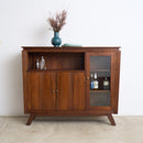 A mid-century modern wooden sideboard cabinet with a warm brown finish, featuring two lower doors, an open shelf, and a glass-front cabinet door, styled with a blue vase on top against a plain white wall.