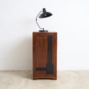 An Art Deco wooden cabinet with a dark brown finish and black inlay details, topped with a black adjustable desk lamp, set against a plain white wall and concrete floor.