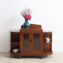A curved Art Deco drinks cabinet with glass-paneled doors and open shelves, styled with a blue vase of pink flowers on top, set against a plain white wall.