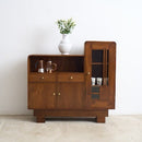 An Art Deco wooden sideboard cabinet with a warm brown finish, featuring a glass-front cabinet on the right, a central drawer, and open shelving on the left. A white ceramic vase with green leaves sits on top.