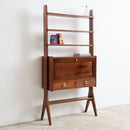 A mid-century modern wooden writing desk with integrated shelves, shown against a plain white wall and light concrete floor.