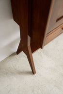 Close-up of a dark brown wooden furniture piece, showing the corner leg and lower drawer area against a white wall and light textured floor.