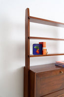 A mid-century modern wooden hutch or shelving unit with a warm brown finish, featuring open shelves and a lower drawer, styled with books and a small decorative tin against a plain white wall.