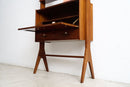 A mid-century modern wooden secretary desk with a warm brown finish, featuring a drop-down writing desk surface, two drawers, and angled legs, set against a plain white wall and concrete floor.