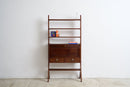 A mid-century modern wooden writing desk with integrated shelving, featuring a dark walnut finish, two drawers, and small decorative objects on the shelves, set against a plain white wall and concrete floor.