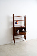 A mid-century modern wooden writing desk and shelving unit with a dark walnut finish, styled with books, a lamp, and small accessories against a white wall and concrete floor.