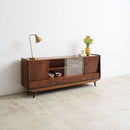 A mid-century modern wooden sideboard cabinet with glass doors and open shelving, styled with a brass table lamp and a small vase of flowers in a minimalist room with a concrete floor and white wall.