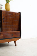 A mid-century modern wooden sideboard cabinet with a warm brown wood finish, featuring a slatted front, drawer, and tapered legs, shown in a minimalist room setting.