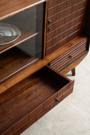 Mid-century modern wooden cabinet with a glass-front upper section, a sliding drawer, and a pull-out storage drawer, shown in warm medium brown wood with dark metal hardware on a light floor.