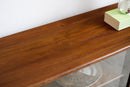 A close-up of a warm brown wooden shelf or tabletop with visible wood grain, photographed indoors against a white wall. A light green book and part of a white mug sit on the surface, and a glass shelf is visible underneath.