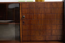 A close-up of a mid-century modern wooden cabinet featuring a mosaic carved teak wood door with a circular metal pull handle and a dark glass side panel.