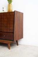 A mid-century modern wooden sideboard cabinet with a dark brown walnut finish, featuring a slatted front, a single drawer with a small round pull, and tapered legs, shown against a white wall.