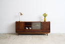 A mid-century modern wooden sideboard cabinet with glass doors and drawers, styled with a brass table lamp and a vase of flowers against a white wall and concrete floor.