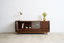 A mid-century modern wooden sideboard cabinet with glass doors and drawers, styled with a brass table lamp and a vase of flowers against a white wall and light stone floor.