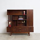 A mid-century modern dark brown wooden hutch with brass nailhead trim, open shelves, two drawers, and brass handles, styled with small decorative objects on a concrete floor against a white wall.