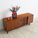 A mid-century modern wooden sideboard cabinet with a warm brown finish, featuring a decorative black and red vase with dried branches on top, set against a plain white wall and light concrete floor.