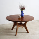 A round teak Vintage dining table with a polished finish and three curved legs, styled with a blue vase of dried flowers and a small bowl on top, set against a minimalist white wall and concrete floor.