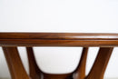 Close-up of a polished medium-brown vintage wooden table top showing the smooth bevelled edge and wood grain, with matching wooden legs blurred in the background against a plain white wall.