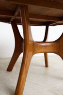 Close-up of a mid-century modern wooden dining table with curved, sculptural legs in a warm medium brown finish, set against a minimalist white wall and light concrete floor.