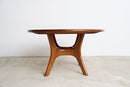 A mid-century modern wooden dining table with a rounded oval top and curved sculptural legs, shown against a plain white wall and light concrete floor.