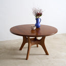 A round wooden coffee table with a mid-century modern design, featuring a dark brown wood finish and curved legs, styled with a small blue vase and dried flowers on a light concrete floor against a white wall.