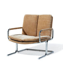 A modernist lounge chair with textured tan fabric upholstery and a polished chrome cantilever frame, shown on a white background.