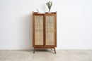 A mid-century modern wooden cabinet with two doors featuring woven cane panels, standing on a light concrete floor against a white wall.