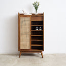 A mid-century modern styled wooden shoe cabinet with a woven cane door panel, open shelves, and tapered legs, shown against a plain white wall and concrete floor.
