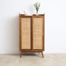 A mid-century modern styled wooden cabinet with two woven cane doors, standing against a white wall on a light concrete floor.
