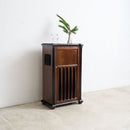 A converted Art Deco wooden speaker cabinet with a dark top and glassware on top, alongside a small potted palm, set against a minimalist white wall and concrete floor.