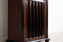 A close-up of a dark brown wooden cabinet or shelf with vertical slatted detailing and black trim, photographed indoors against a plain light wall.