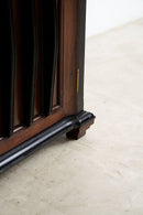 Close-up detail of a dark brown wooden cabinet corner with a black metal guard rail and turned wooden feet, set on a light beige stone floor.