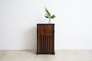 A dark brown wooden cabinet with a slatted front door and small brass knobs, standing against a plain white wall on a light concrete floor. A small potted palm and a glass are placed on top of the cabinet.