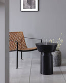 Black Nanded side table placed in front of a woven chair. On top of the table there is a glass placed on its top.