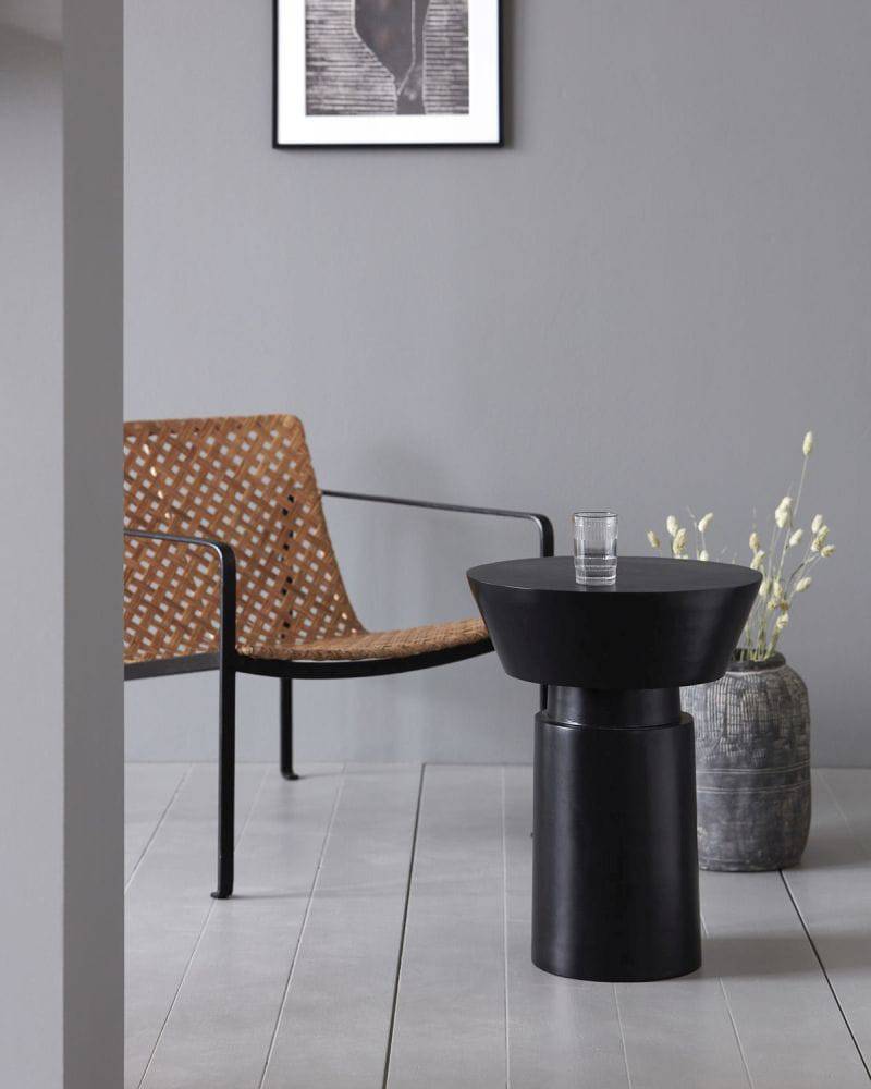 Black Nanded side table placed in front of a woven chair. On top of the table there is a glass placed on its top.